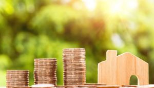 Three stacks of coins to the left of a small wood block shaped like a house with a blurred green background.